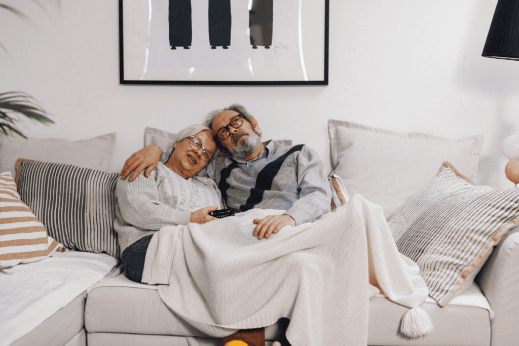 An elderly couple sits on a cozy couch, snuggled under a blanket. Smiling and relaxed with a TV remote, they feel confident thanks to discreet bladder leak pads. Framed art and pillows decorate the background.