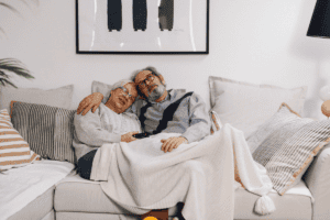 An elderly couple sits on a cozy couch, snuggled under a blanket. Smiling and relaxed with a TV remote, they feel confident thanks to discreet bladder leak pads. Framed art and pillows decorate the background.