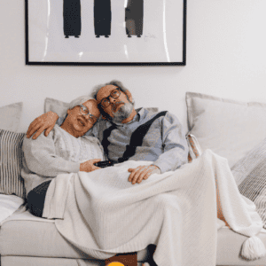 An elderly couple sits on a cozy couch, snuggled under a blanket. Smiling and relaxed with a TV remote, they feel confident thanks to discreet bladder leak pads. Framed art and pillows decorate the background.
