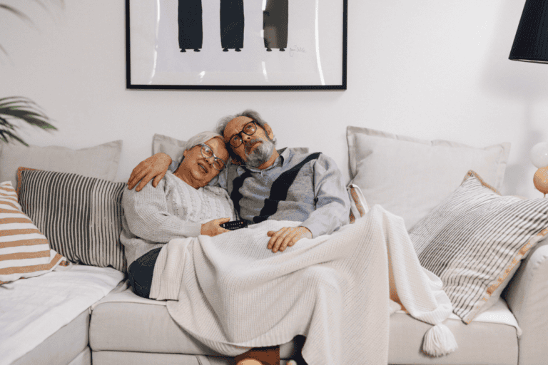 An elderly couple sits on a cozy couch, snuggled under a blanket. Smiling and relaxed with a TV remote, they feel confident thanks to discreet bladder leak pads. Framed art and pillows decorate the background.