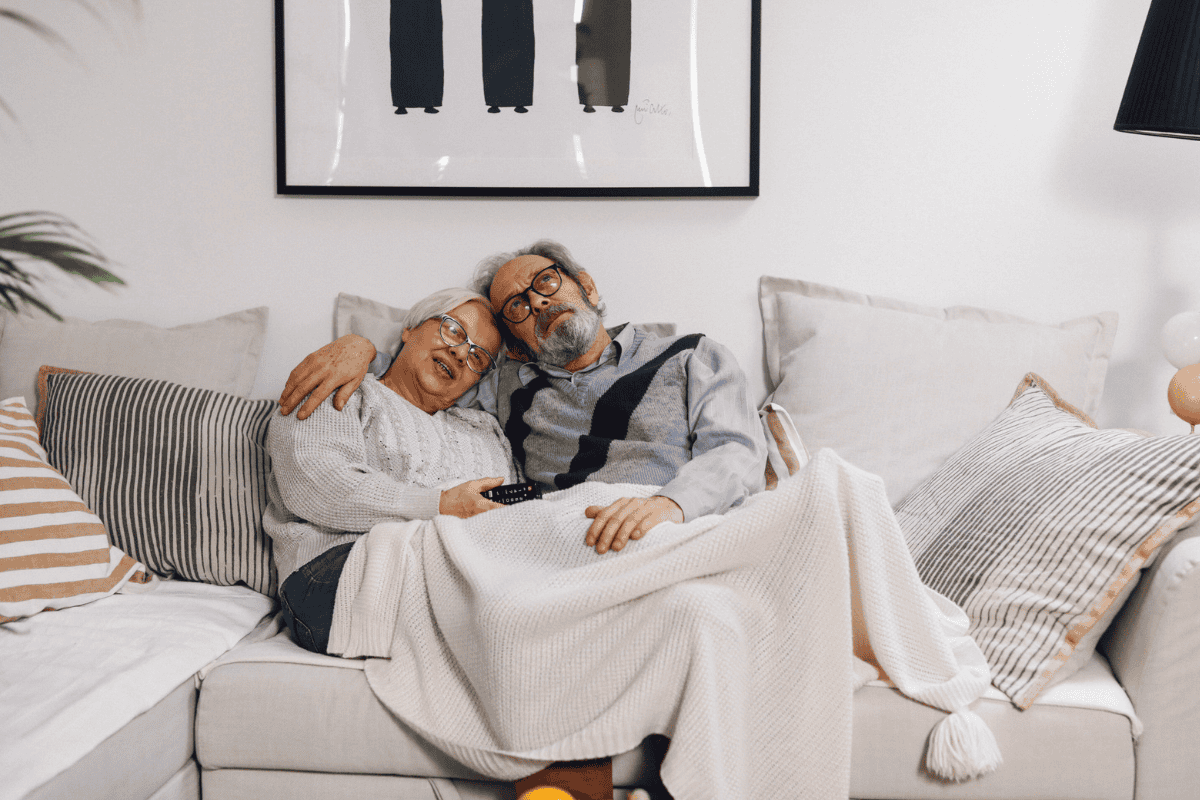 ATETranquilityBlog2026 An elderly couple sits on a cozy couch, snuggled under a blanket. Smiling and relaxed with a TV remote, they feel confident thanks to discreet bladder leak pads. Framed art and pillows decorate the background.