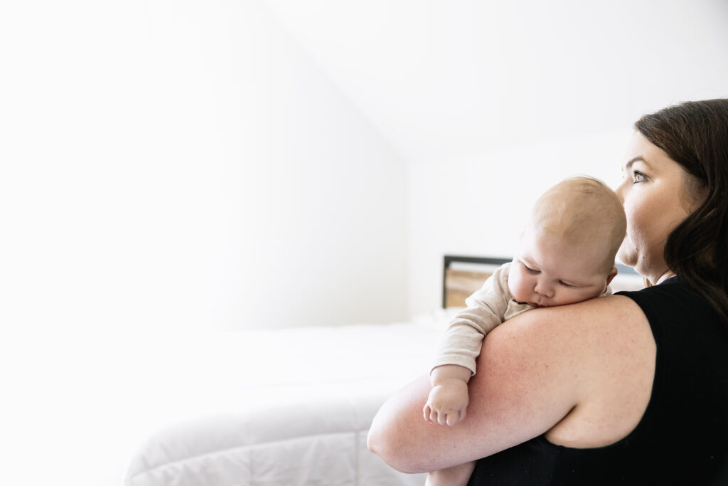 A woman with long brown hair holds a baby resting on her shoulder in a bright, white bedroom. Both are facing away from the camera toward the light from a window, reflecting quiet moments after pregnancy and recovery from prolapse after pregnancy.