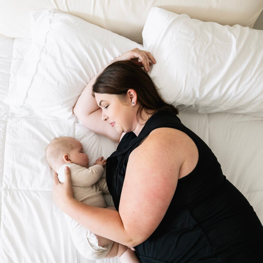 A woman in a black dress lies on a white bed, cradling a sleeping baby in light-colored clothes. Both rest peacefully, with the woman looking lovingly at the baby—capturing tender moments after pregnancy, when concerns like prolapse after pregnancy may arise.
