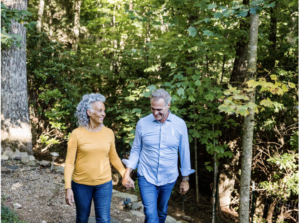 An older couple walks hand in hand on a forest path, smiling at each other. Sunlight filters through green trees, creating a peaceful, natural setting. Thanks to reliable bladder leak pads, they enjoy their stroll with confidence and comfort.