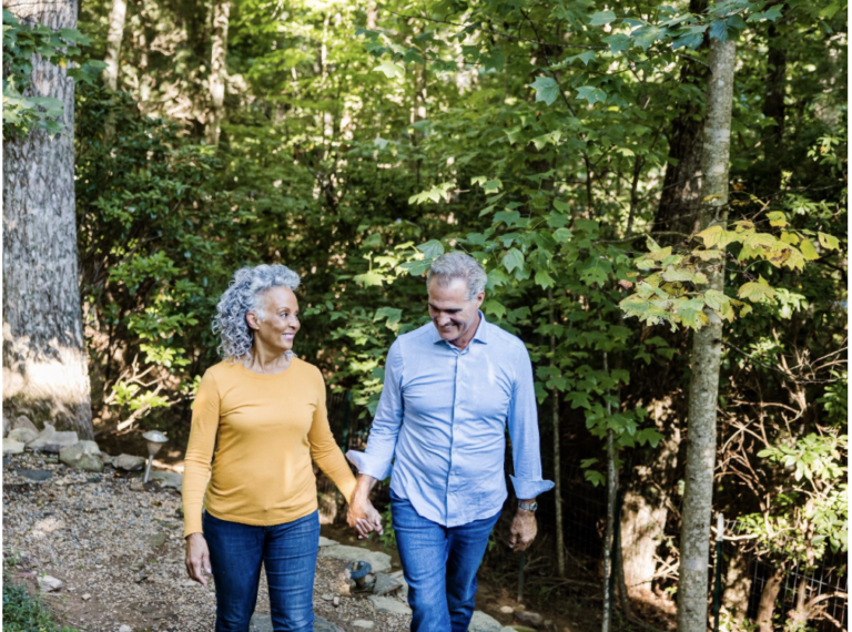 An older couple walks hand in hand on a forest path, smiling at each other. Sunlight filters through green trees, creating a peaceful, natural setting. Thanks to reliable bladder leak pads, they enjoy their stroll with confidence and comfort.