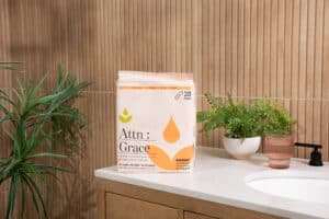 A pack labeled Attn: Grace sits on a bathroom counter next to a pink cup, potted plants, and a black faucet—an inviting scene highlighting support for incontinence after prostate cancer surgery amid light wood paneling and a white sink.