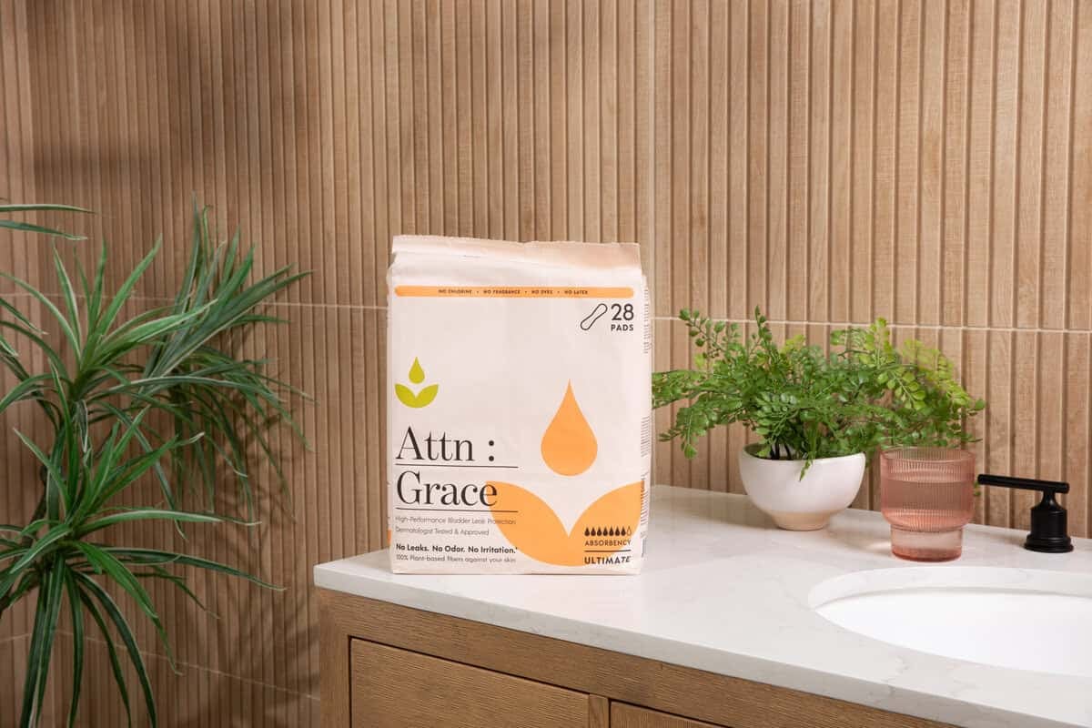 Attn Grace_r2_051A1456-ynyhwl1715 A pack labeled Attn: Grace sits on a bathroom counter next to a pink cup, potted plants, and a black faucet—an inviting scene highlighting support for incontinence after prostate cancer surgery amid light wood paneling and a white sink.