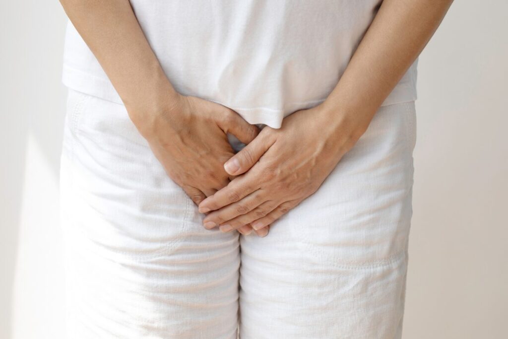 A person dressed in white clothes stands with their hands covering their lower abdomen and groin area, possibly indicating discomfort or pain from incontinence skin irritation, a frequent symptom seen in common types of urinary incontinence.