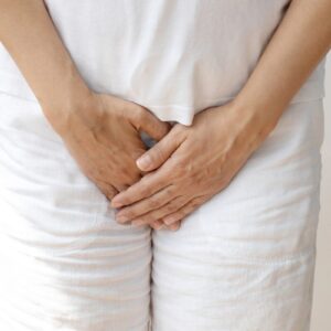 A person dressed in white clothes stands with their hands covering their lower abdomen and groin area, possibly indicating discomfort or pain from incontinence skin irritation, a frequent symptom seen in common types of urinary incontinence.