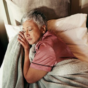 An older woman with short gray hair sleeps peacefully in bed, wearing a pink satin pajama top and hugging a soft gray blanket. A glass of water, medicine bottles, and items to help manage incontinence cost sit on the nightstand beside her.
