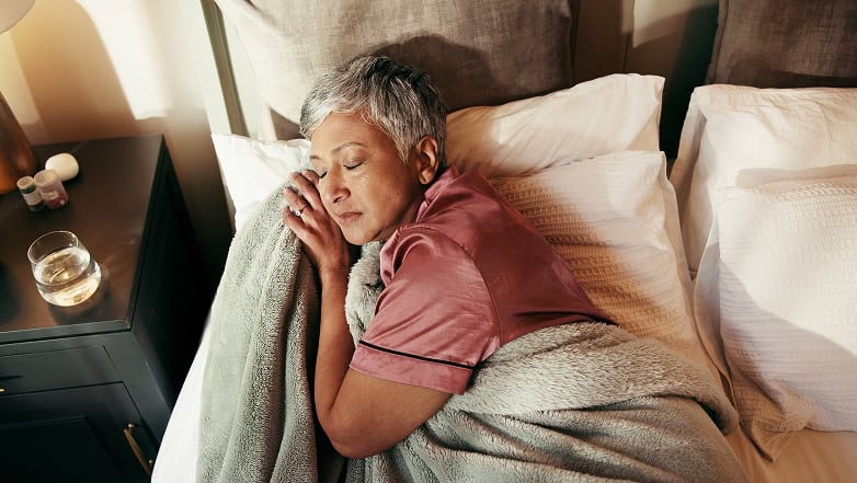 Header_Image_2 An older woman with short gray hair sleeps peacefully in bed, wearing a pink satin pajama top and hugging a soft gray blanket. A glass of water, medicine bottles, and items to help manage incontinence cost sit on the nightstand beside her.