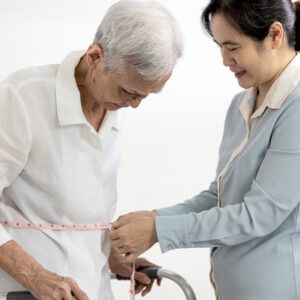 A smiling caregiver measures the waist of an elderly person using a measuring tape. The elderly person is standing with the aid of a walker. Both are indoors, and the atmosphere appears supportive and caring.