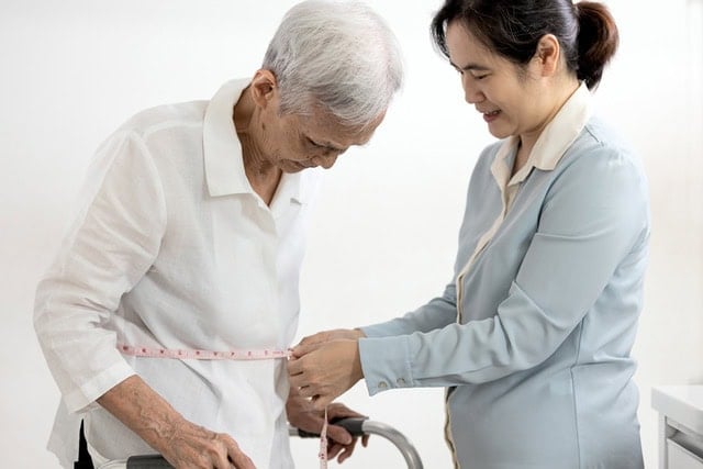 Measuring Waist A smiling caregiver measures the waist of an elderly person using a measuring tape. The elderly person is standing with the aid of a walker. Both are indoors, and the atmosphere appears supportive and caring.
