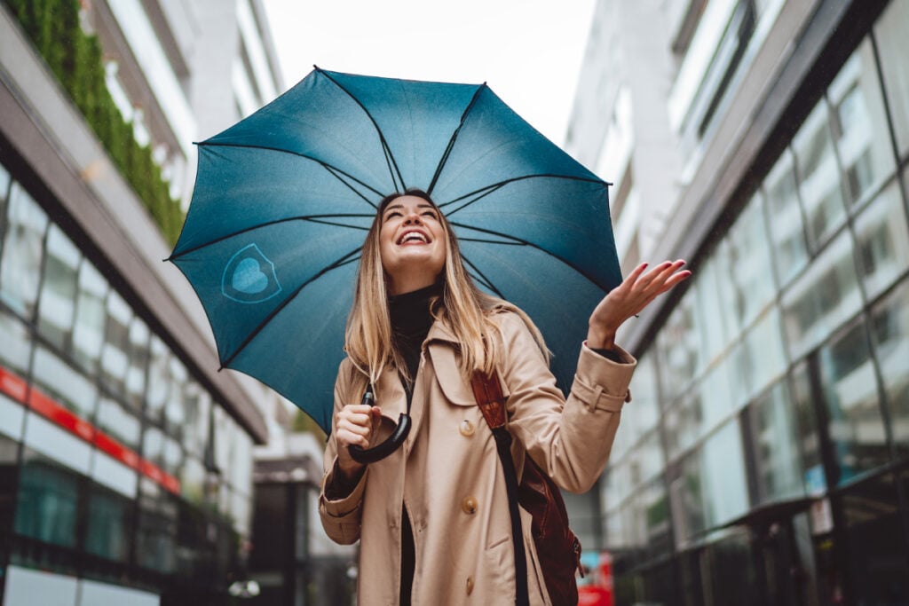 A woman in a beige coat smiles and looks up while holding a blue umbrella on a city street, surrounded by modern glass buildings—a perfect scene to reflect on how to improve gut health and embrace the secrets of your gut for overall well-being.
