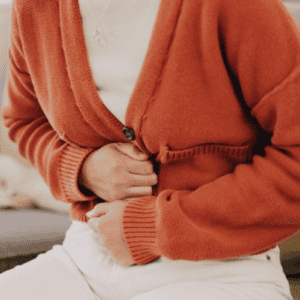 A person wearing an orange cardigan and white pants sits on a couch, clutching their stomach with both hands, appearing to be in discomfort or pain—perhaps a sign that attending to gut health is more important than ever.
