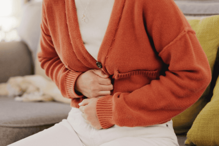 A person wearing an orange cardigan and white pants sits on a couch, clutching their stomach with both hands, appearing to be in discomfort or pain—perhaps a sign that attending to gut health is more important than ever.