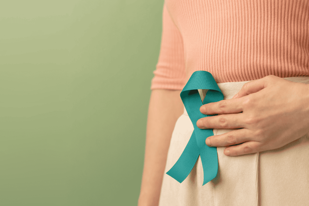 A person wearing a peach top and beige skirt holds a teal awareness ribbon pinned to their waist, standing against a light green background—highlighting the importance of gut health and raising awareness about how to improve gut health.