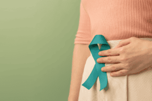 A person wearing a peach top and beige skirt holds a teal awareness ribbon pinned to their waist, standing against a light green background—highlighting the importance of gut health and raising awareness about how to improve gut health.