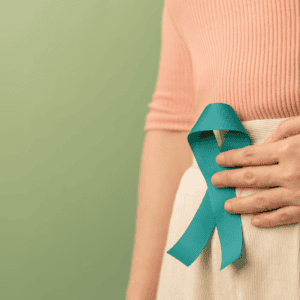 A person wearing a peach top and beige skirt holds a teal awareness ribbon pinned to their waist, standing against a light green background—highlighting the importance of gut health and raising awareness about how to improve gut health.