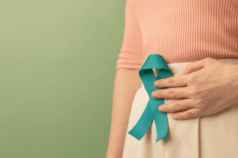 A person wearing a peach top and beige skirt holds a teal awareness ribbon pinned to their waist, standing against a light green background—highlighting the importance of gut health and raising awareness about how to improve gut health.