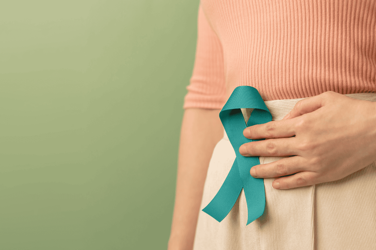 NAFCBladderCancerBlog2026 A person wearing a peach top and beige skirt holds a teal awareness ribbon pinned to their waist, standing against a light green background—highlighting the importance of gut health and raising awareness about how to improve gut health.