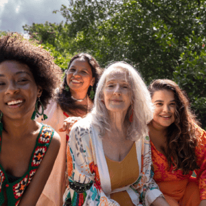 Four women of different ages sit closely together outdoors by water and greenery, smiling at the camera. The group appears joyful and relaxed, confidently enjoying the sunny day thanks to absorbent products for bladder leakage in women.