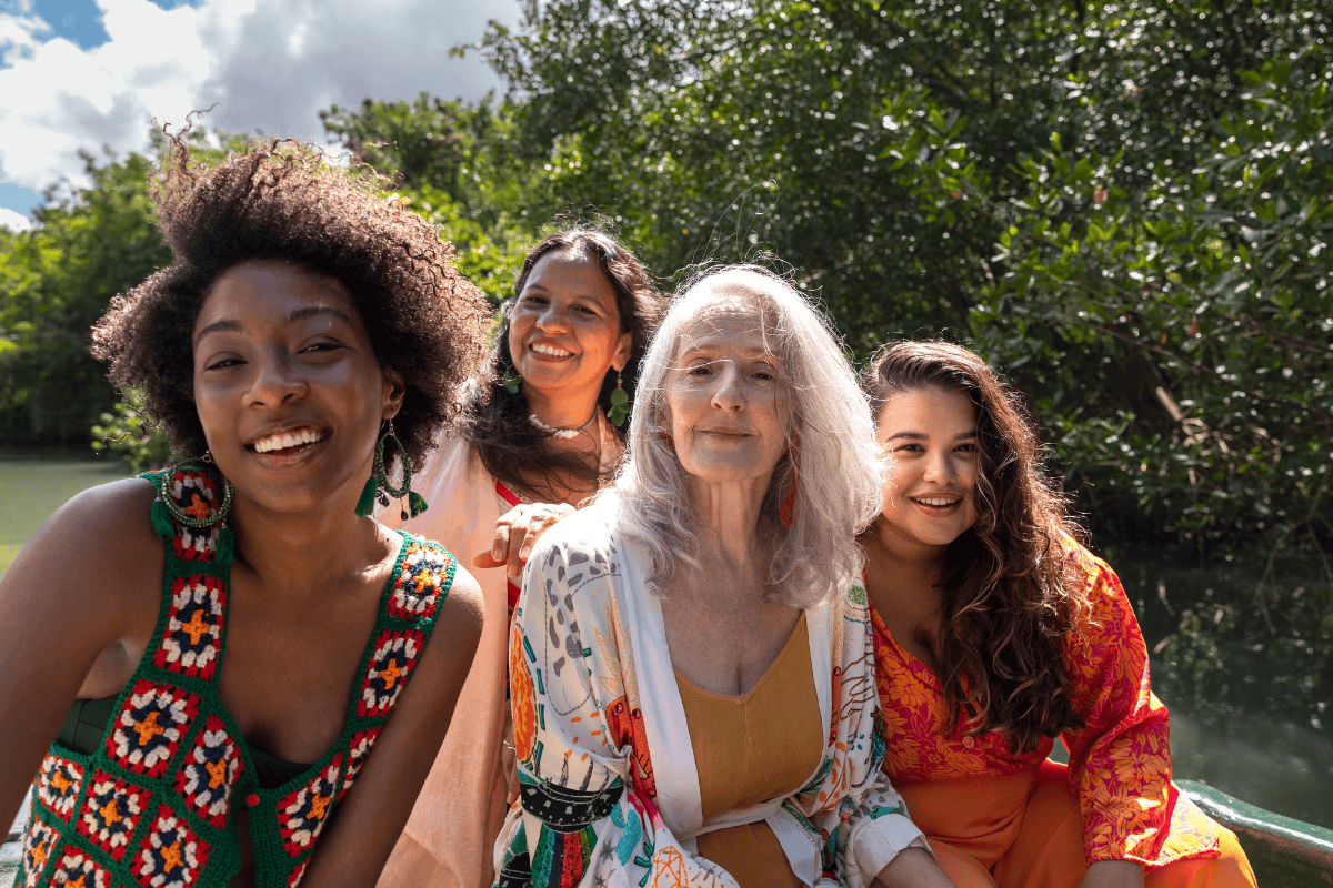 NAFCPutYourselfFirst2026 Four women of different ages sit closely together outdoors by water and greenery, smiling at the camera. The group appears joyful and relaxed, confidently enjoying the sunny day thanks to absorbent products for bladder leakage in women.