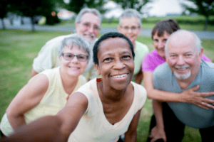 A group of six smiling adults takes a selfie outdoors in a grassy area, with trees and a road in the background. Everyone appears happy and relaxed, enjoying each other’s company—there’s no sign anyone is worried about a UTI, just laughter and good times shared among friends.