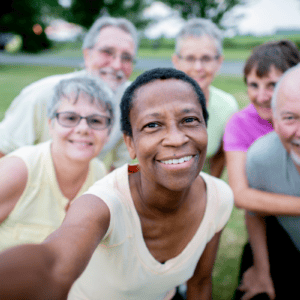 A group of six smiling adults takes a selfie outdoors in a grassy area, with trees and a road in the background. Everyone appears happy and relaxed, enjoying each other’s company—there’s no sign anyone is worried about a UTI, just laughter and good times shared among friends.