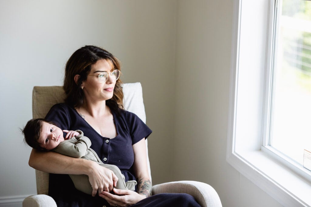 A woman with glasses sits in a beige armchair holding her baby, both looking out a bright window. The peaceful, light-filled room offers comfort as she gently cares for her newborn while recovering from prolapse after pregnancy.