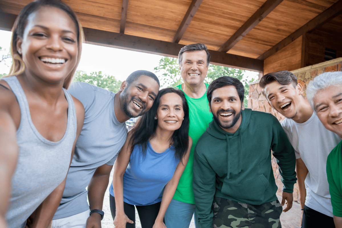 A group of seven diverse adults, including one holding a pamphlet about UTI awareness, smiling and posing closely together for a selfie outside under a wooden roof.
