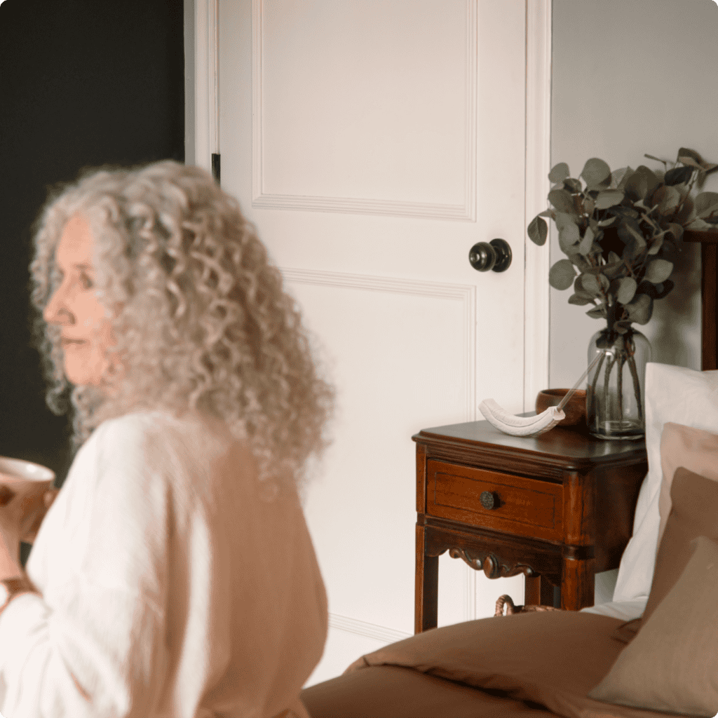 An older woman with curly gray hair sits on a bed holding a cup. A wooden nightstand with greenery and a white ceramic dish is beside her, next to a white door. The calm, cozy scene highlights comfort—a focus of Featured Partner: Aeroflow Urology.
